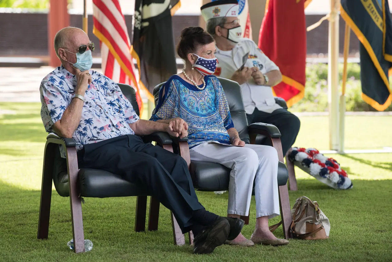 Jim and Carole Hickerson seated side by side at an outdoor ceremony, holding hands, with flags behind them.